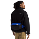 A man wearing a black fleece jacket and khaki pants is shown from the back, wearing a black and blue waist bag across his lower back. The brand 'The North Face' is visible on the jacket's upper back and the bag.