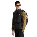 A model wearing a black puffer vest with a front zipper and buckle closure, layered over an olive green long-sleeve shirt, black pants, and a tan backpack, set against a plain white background.