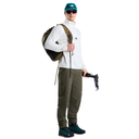 A model wearing a white long-sleeve technical top, olive green pants, a green and black backpack, and a green cap, holding a pair of ski boots against a solid white background.
