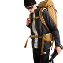 A man wearing a black beanie, sunglasses, a black jacket, and black pants models a tan and brown technical hiking backpack with a hip belt and shoulder harness system. The The North Face logo is visible on his pants.