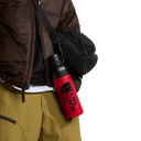 A red and black The North Face water bottle with a black carabiner clip, hanging from a black crossbody bag worn by a person in a brown puffer jacket and olive pants, against a white background.
