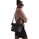 A woman wearing a tan jacket, black shorts, and sunglasses carries a black crossbody bag from The North Face on a plain white background.