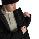 A close-up of a person wearing a black jacket over a light beige henley shirt and a black knit beanie, with their hands opening the jacket zipper, against a solid white background.