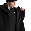A man wearing a black The North Face jacket over a black puffer vest and a black beanie, looking down while holding the jacket's collar open against a white background.