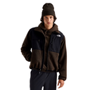 A male model wearing a brown and black fleece jacket with a hood, a matching brown beanie, and dark pants, photographed against a solid white background. The jacket features a brown sherpa-lined front and a black outer shell with zippered pockets.