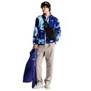 A model stands against a white background wearing a blue and white tie-dye fleece jacket, beige pants, and white sneakers, while holding a blue puffer jacket and wearing a black crossbody bag.