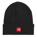 A black ribbed knit beanie hat with a folded cuff, featuring a small red square patch on the front with the white text and logo of The North Face.