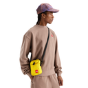 A model wearing a beige sweatshirt and matching pants, a purple patterned cap, and a bright yellow crossbody bag with a small red logo, shown against a plain white background.
