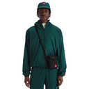 A model wearing a dark green hooded windbreaker jacket, matching green pants, a green cap, and a black crossbody bag with a red logo patch, shot against a solid white background.