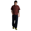 A model stands against a solid white background wearing a red and black plaid short-sleeve button-up shirt, dark blue jeans, and lime green sneakers.
