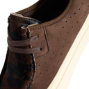 Close-up of a brown suede shoe with perforated detailing, matching brown laces, and a dark brown and black patterned fur trim along the collar, set against a white background.