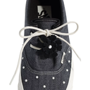 A close-up of a dark gray canvas sneaker featuring white laces tied in a bow with a black fabric flower accent, white studs, and a white rubber sole.
