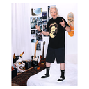 A man stands on a white bed in a bedroom, wearing a black graphic T-shirt with a large circular face print, black shorts, black socks, and black sneakers. The room has guitars, a skateboard, posters, a horned mask, and other bedroom decor visible.