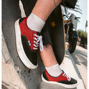 A person wearing red and black low-top sneakers with white socks, sitting on concrete outdoors with a skateboard and a motorcycle in the background.