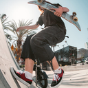 A skateboarder in a black t-shirt and black denim shorts rides a skateboard while balancing on a concrete ramp, with palm trees and a sunny outdoor city background.