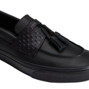 A close-up of a black leather slip-on loafer with a textured woven strap and a black tassel detail, set against a plain white background.
