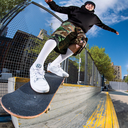A skateboarder in a black hoodie, camouflage shorts, white socks, and white sneakers performs a trick on a concrete ledge in an urban outdoor setting under a blue sky.