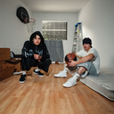 Two young men posing in a room with a wooden floor, basketball hoop, and mattress. One is wearing a black hoodie with blue graphics, black pants, and black/white sneakers, while the other wears a white t-shirt, gray shorts, white sneakers, and a black beanie, holding a basketball.