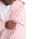 A close-up of a person wearing a light pink zip-up hoodie over a white t-shirt. The hoodie features a black 'ATIBA > VANS' graphic printed on the left sleeve pocket.