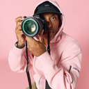 Atiba wearing a light pink hoodie and a black Vans cap holds a black Canon EOS camera up to their eye, photographed against a solid pink studio background.