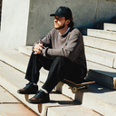 A man sits on concrete stairs wearing a dark baseball cap, a gray crewneck sweater, black pants, and dark brown skate shoes, with a skateboard resting on the step beside him.