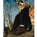 A person sitting outdoors on a stone ledge wearing brown suede high-top skate shoes, black pants, and a brown jacket, with bare trees and a colorful mosaic wall in the background.