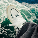 A white low-top sneaker with white laces and black trim, worn with dark gray jeans, photographed with foamy green-blue water in the background.
