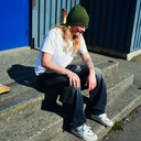 A person sitting on concrete steps outdoors, wearing a green beanie, white graphic T-shirt, dark jeans, and white sneakers, with a blue metal wall in the background.