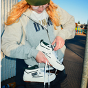 A person outdoors wearing a gray hoodie and green beanie while holding and lacing up a pair of white low-top sneakers with black and green accents. The sneakers have a visible 'KORS' logo patch on the side.