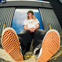 A person sitting outdoors in front of a corrugated metal wall and blue door, wearing a white graphic T-shirt, dark jeans, a green beanie, and orange-brown Vans sneakers. The photo is taken from a low angle with the soles of both shoes prominently visible.
